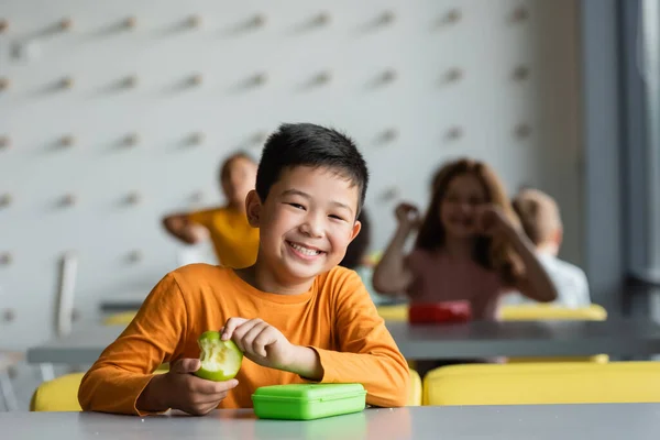Счастливый азиатский ребенок держит со свежим яблоком улыбаясь в камеру возле размытых детей в школьной столовой — стоковое фото