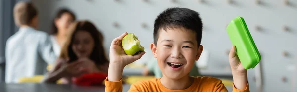 Cheerful asian boy holding lunch box and apple near blurred kids in school eatery, banner — Stock Photo