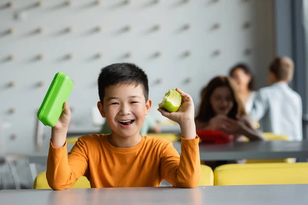 Alegre asiático chico celebración almuerzo caja y manzana en escuela comedor - foto de stock