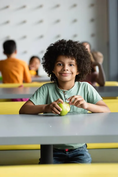 Афріканський американський школяр з свіжим яблуком, який посміхається біля розмитих дітей. — стокове фото