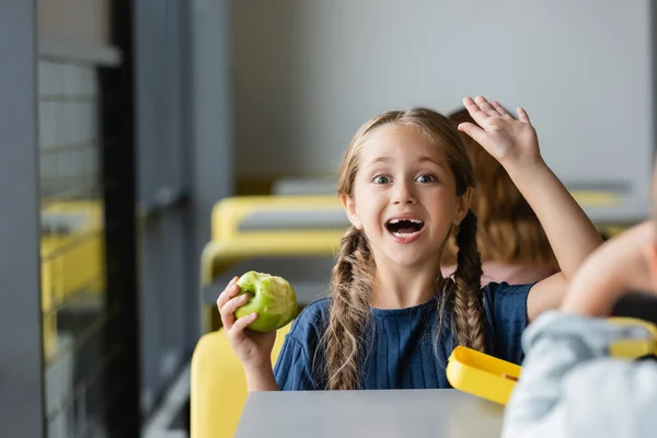 Excitada chica sosteniendo manzana y saludando la mano en el comedor de la escuela - foto de stock