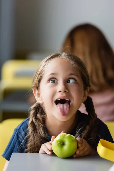 Ragazza giocosa con smorfie di mele durante la pausa pranzo nella sala da pranzo della scuola — Foto stock