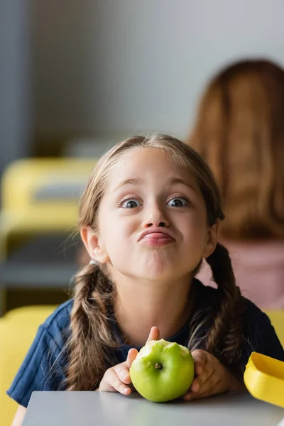 Juguetona chica sobresaliendo lengua mientras mueca cerca de manzana en la cantina de la escuela - foto de stock