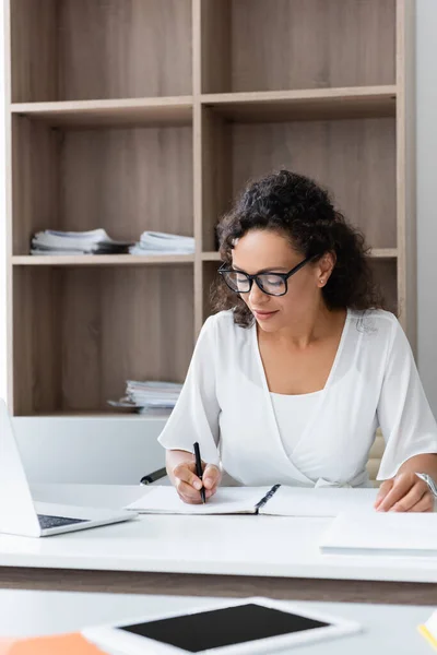 African american teacher in eyeglasses writing in notebook near laptop — Stock Photo