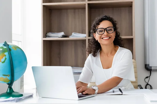 Glücklich afrikanisch-amerikanischer Lehrer lächelt in die Kamera, während er auf dem Laptop im Klassenzimmer tippt — Stockfoto