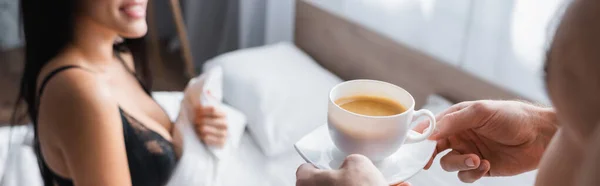 Cropped view of blurred and shirtless man holding cup of cappuccino near seductive woman in underwear in bedroom, banner — Stock Photo