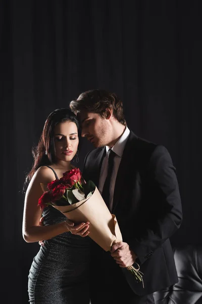 Young man in suit holding bouquet of red roses near girlfriend in elegant dress on black — Stock Photo