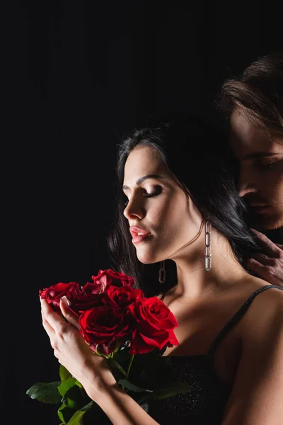 Giovane donna che tiene bouquet di rose rosse vicino all'uomo isolato su nero — Foto stock