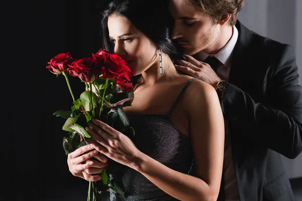 Young woman smelling bouquet of red roses near man in suit on black — Stock Photo