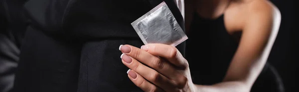 Обрізаний вид жінки, що тримає презерватив, маскуючись з чоловіком на чорному, банер — стокове фото