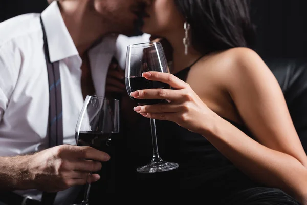 Vista cortada de casal borrado e sexy segurando copos de vinho tinto enquanto se beija isolado no preto — Fotografia de Stock