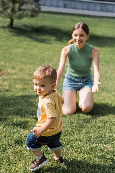 Blurred cheerful mother with outstretched hands sitting on grass and reaching toddler son outside — Stock Photo