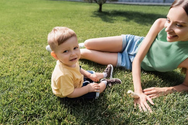 Glückliche junge Mutter schaut fröhlichen Kleinkind-Sohn auf Gras an — Stockfoto