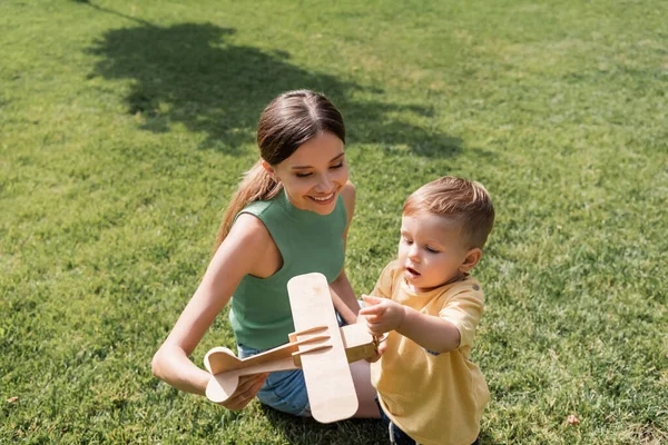 Lächelnde Mutter hält Spielzeug-Doppeldecker neben Kleinkind-Sohn im Gras — Stockfoto