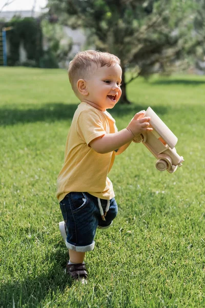 Счастливый мальчик в полный рост с игрушечной деревянной машиной, стоящей на зеленой траве — стоковое фото
