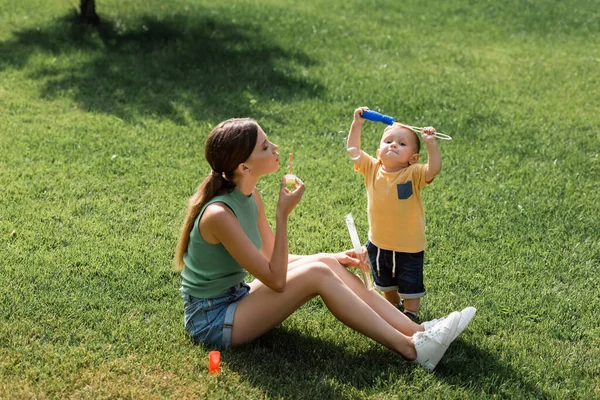 Young mother sitting on grass and blowing soap bubbles near toddler son — Stock Photo