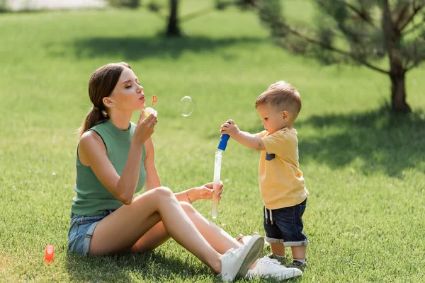 Madre seduta sull'erba e soffiando bolla di sapone vicino al bambino ragazzo — Foto stock