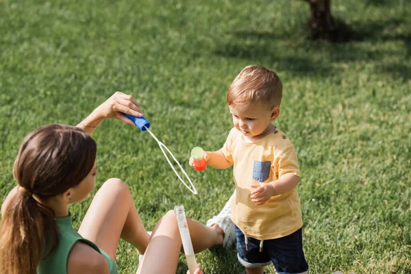 Borrosa madre sosteniendo burbuja varita cerca de niño pequeño hijo fuera - foto de stock