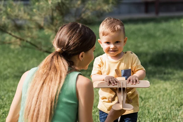 Mère regardant tout-petit garçon avec biplan en bois à l'extérieur — Photo de stock