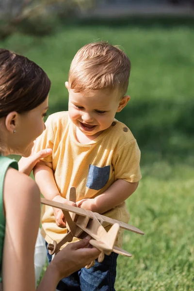 Blurred cheerful mother near smiling toddler boy with wooden biplane — Stock Photo