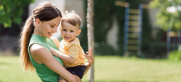 Fröhliche und junge Mutter hält Kleinkind Sohn in den Armen herausstreckende Zunge, Banner — Stockfoto