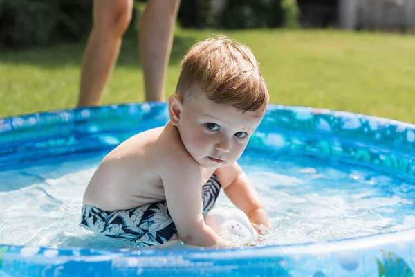 Niño en traje de baño sentado en la piscina inflable cerca de la madre - foto de stock