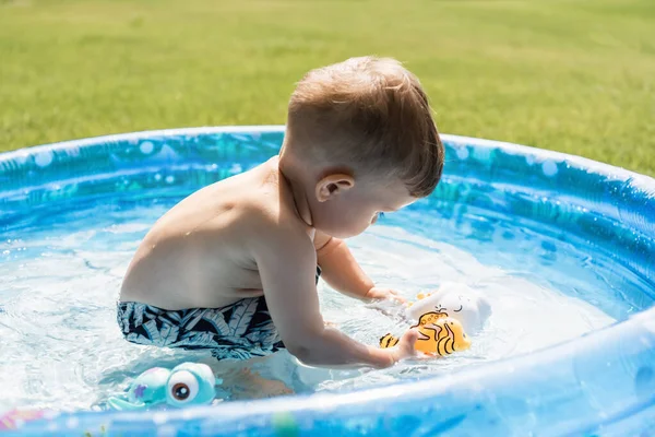 Toddler boy in swim trunks playing with rubber toys in inflatable pool outside — Stock Photo