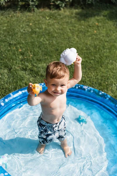 High angle view of happy toddler boy in swim trunks playing with rubber toys in inflatable pool outside — Stock Photo