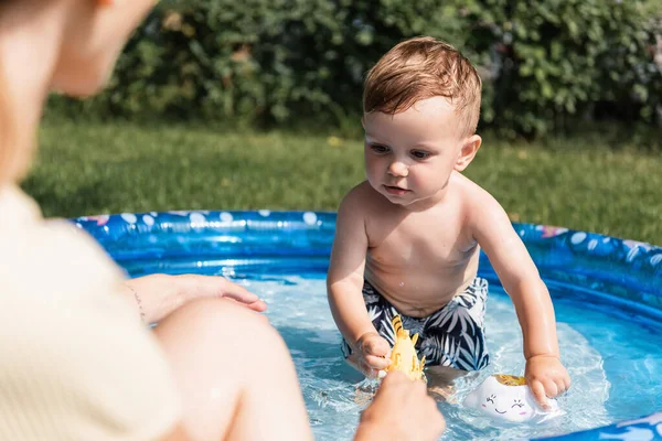 Niño pequeño en baúles de natación jugando con juguetes de goma en piscina inflable cerca de madre borrosa - foto de stock