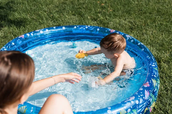 Високий кут зору хлопчика, який грає з гумовими іграшками в надувному басейні поблизу розмитої матері — стокове фото
