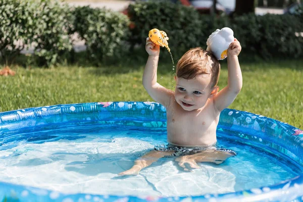 Niño alegre jugando con juguetes de goma y vertiendo agua en la piscina inflable al aire libre - foto de stock