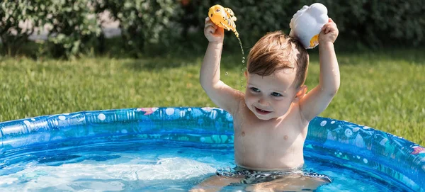 Cheerful toddler boy playing with rubber toys and pouring water in inflatable pool outside, banner — Stock Photo