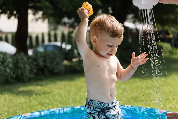 Cheerful toddler boy playing with rubber toy near pouring water outside — Stock Photo