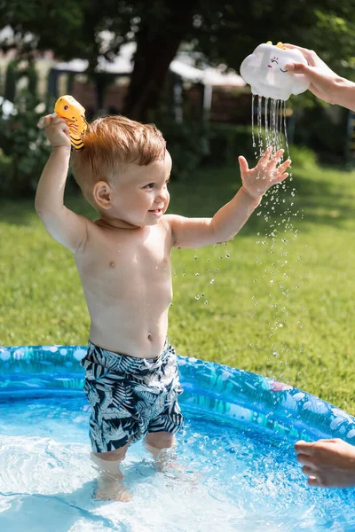 Happy toddler boy in swim trunks near mother holding rubber toy and pouring water outside — Stock Photo