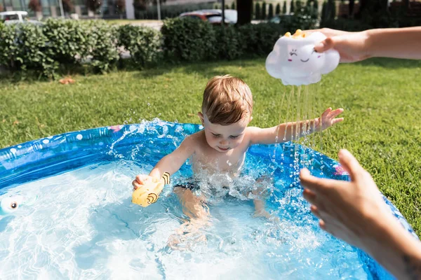 Toddler boy sitting in inflatable pool near mother pouring water from rubber toy outside — Stock Photo