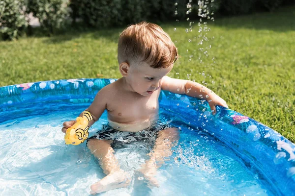 Bambino bambino seduto in piscina gonfiabile con giocattolo di gomma all'esterno — Foto stock