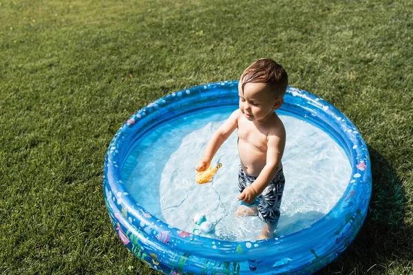 High angle view of cheerful toddler boy standing in inflatable pool with rubber toy — Stock Photo
