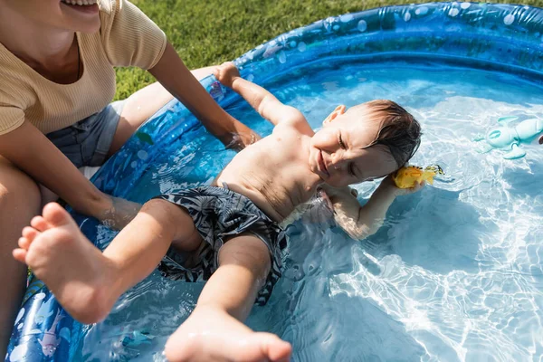 Cheerful mother supporting toddler son swimming in inflatable pool — Stock Photo
