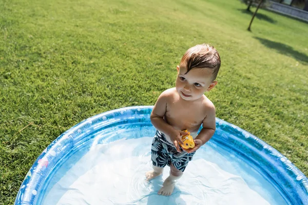 High angle view of toddler child in swim trunks standing in inflatable pool with rubber toy — Stock Photo