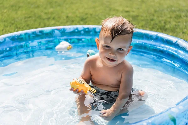 Niño positivo sentado en la piscina inflable y sosteniendo el juguete de goma - foto de stock