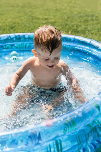 Niño sentado en la piscina inflable y haciendo salpicaduras de agua - foto de stock