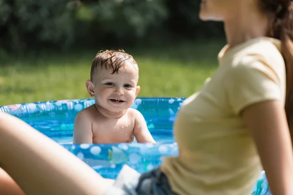 Positivo bambino ragazzo seduto in piscina gonfiabile vicino alla madre offuscata — Foto stock