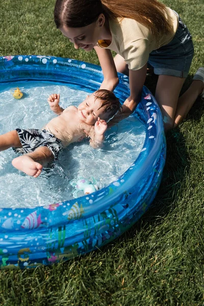 Vista de ángulo alto del hijo del niño del baño de la madre feliz en la piscina inflable - foto de stock