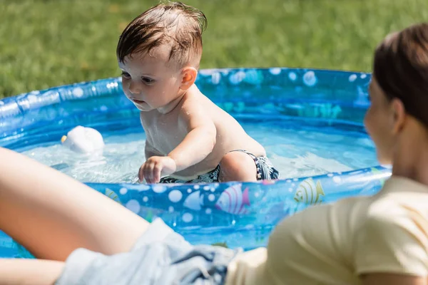 Niño pequeño sentado en la piscina inflable cerca de la madre borrosa en primer plano - foto de stock
