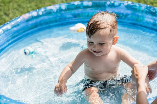 Madre cerca sonriente hijo sentado en piscina inflable con agua azul - foto de stock