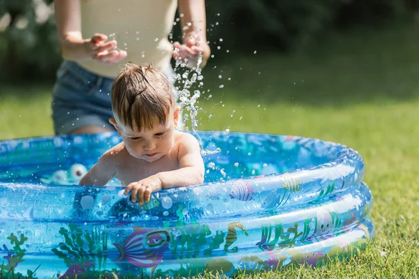 Madre vertiendo agua sobre hijo sentado en piscina inflable - foto de stock