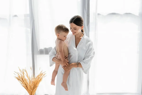 Cheerful mother in bathrobe holding in arms naked toddler son — Stock Photo