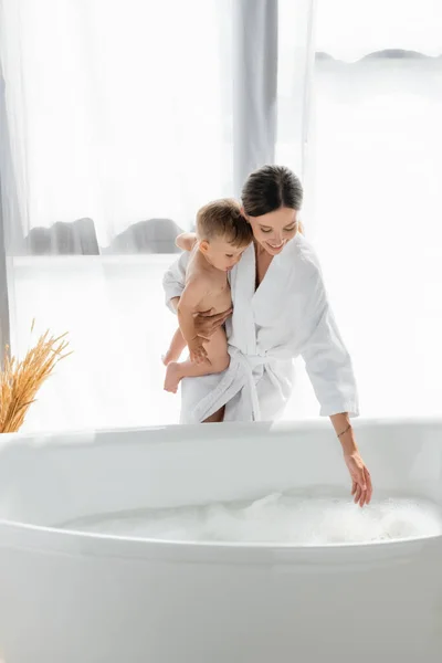 Caring mother in bathrobe holding in arms naked toddler son near bathtub with bath foam — Stock Photo