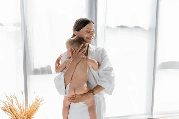 Smiling mother in bathrobe holding in arms naked toddler son — Stock Photo