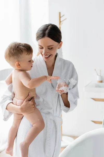 Alegre madre en albornoz sosteniendo en brazos hijo pequeño cerca de la bañera con espuma de baño - foto de stock